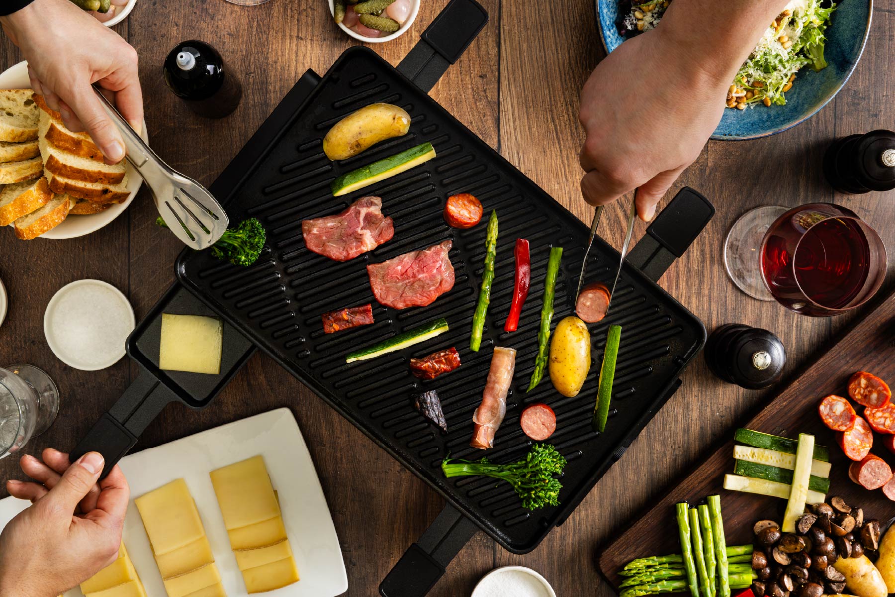 Grilling vegetables and meats on a tabletop grill with hands using tongs.