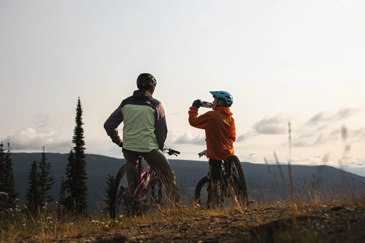 Two cyclists at a scenic overlook, one drinking water, with mountains in the background.