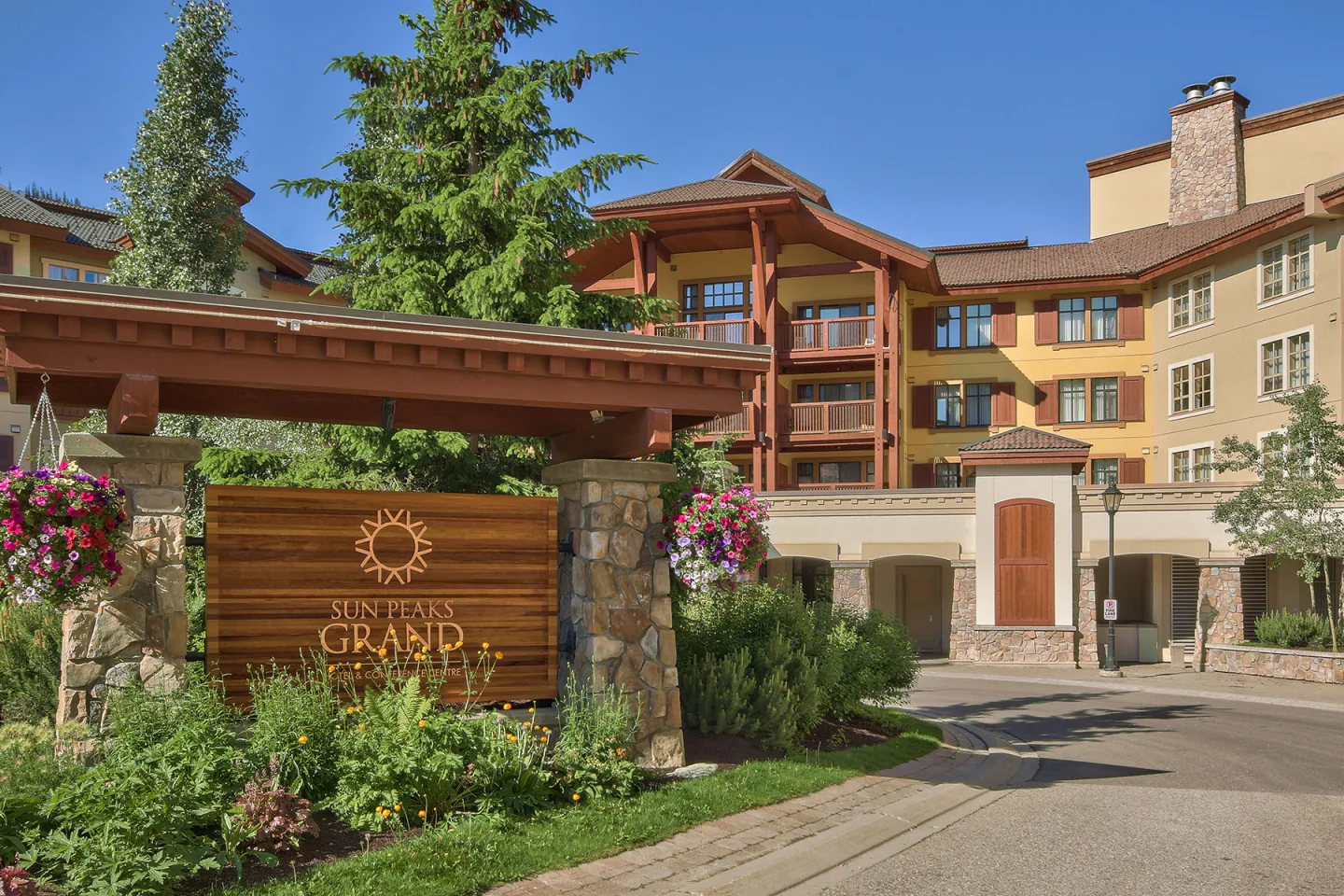 Hotel entrance with wooden sign, colorful flowers, and sunny sky.