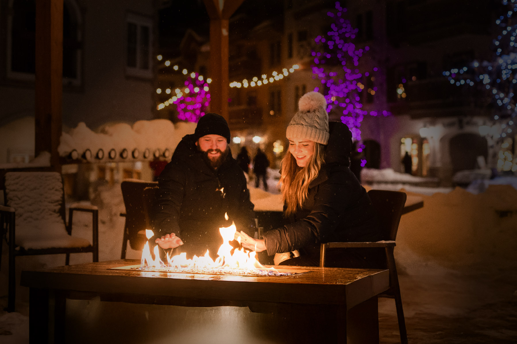 Two people in winter attire gather around a glowing outdoor fire pit at night.