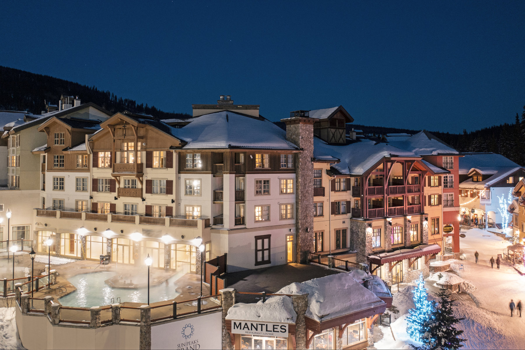 Snowy resort at night, warmly lit buildings, mountain backdrop.
