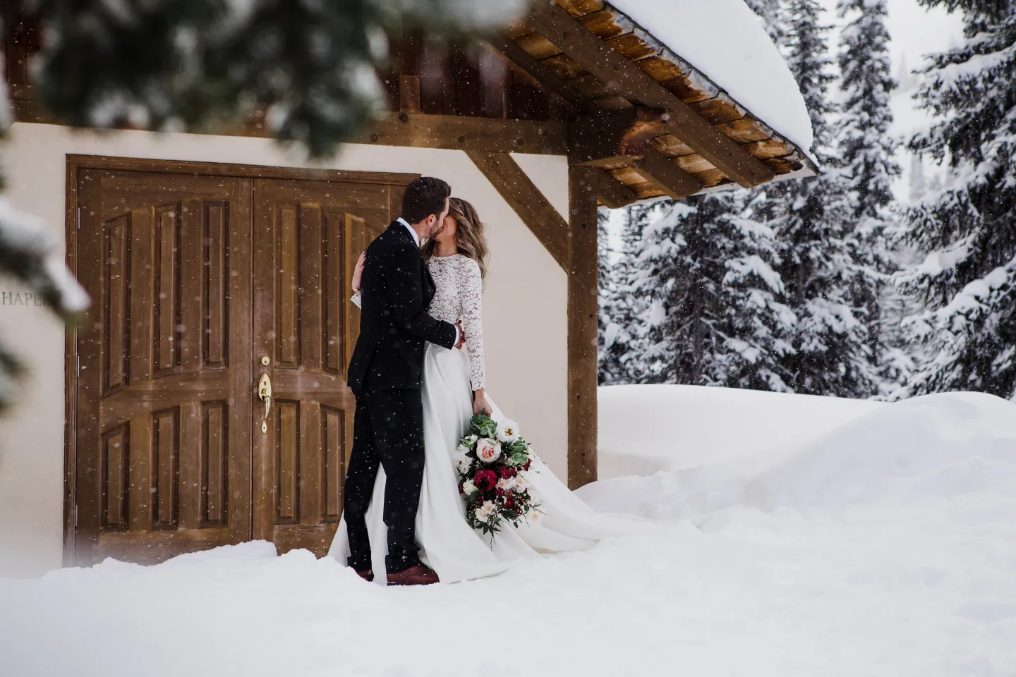 A bride and groom kiss beneath a wooden chapel outside in the snow