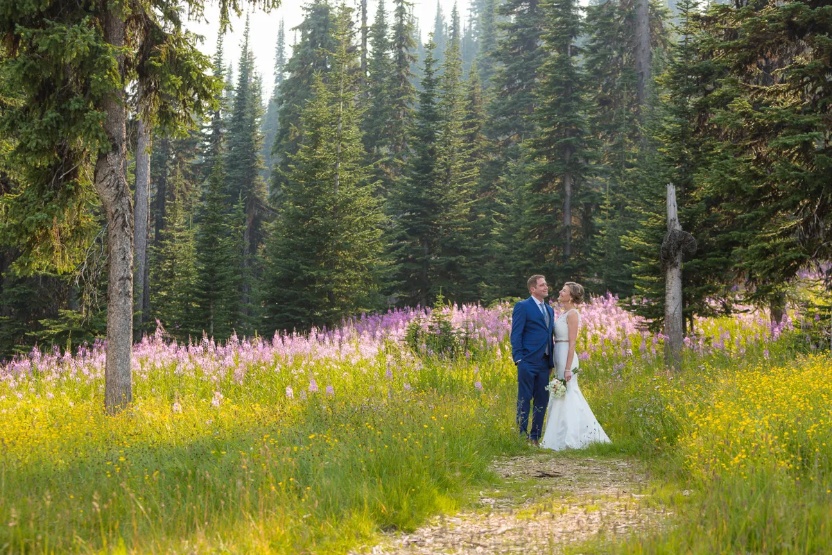 A bride and a groom stand in a meadow surrounded by trees and flowers