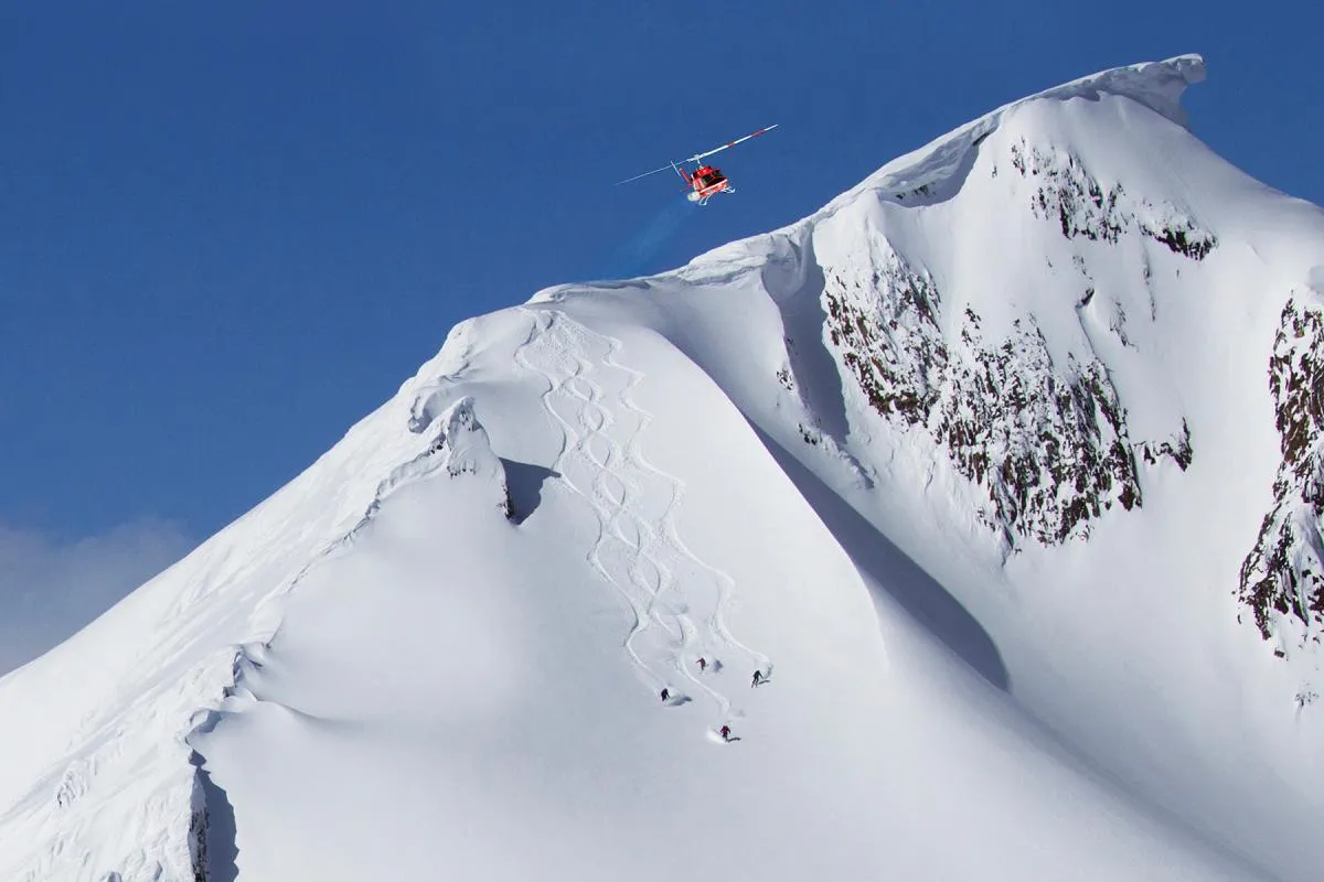 A view of a snowy mountain with four skiers skiing down a face with a helicopter in the background