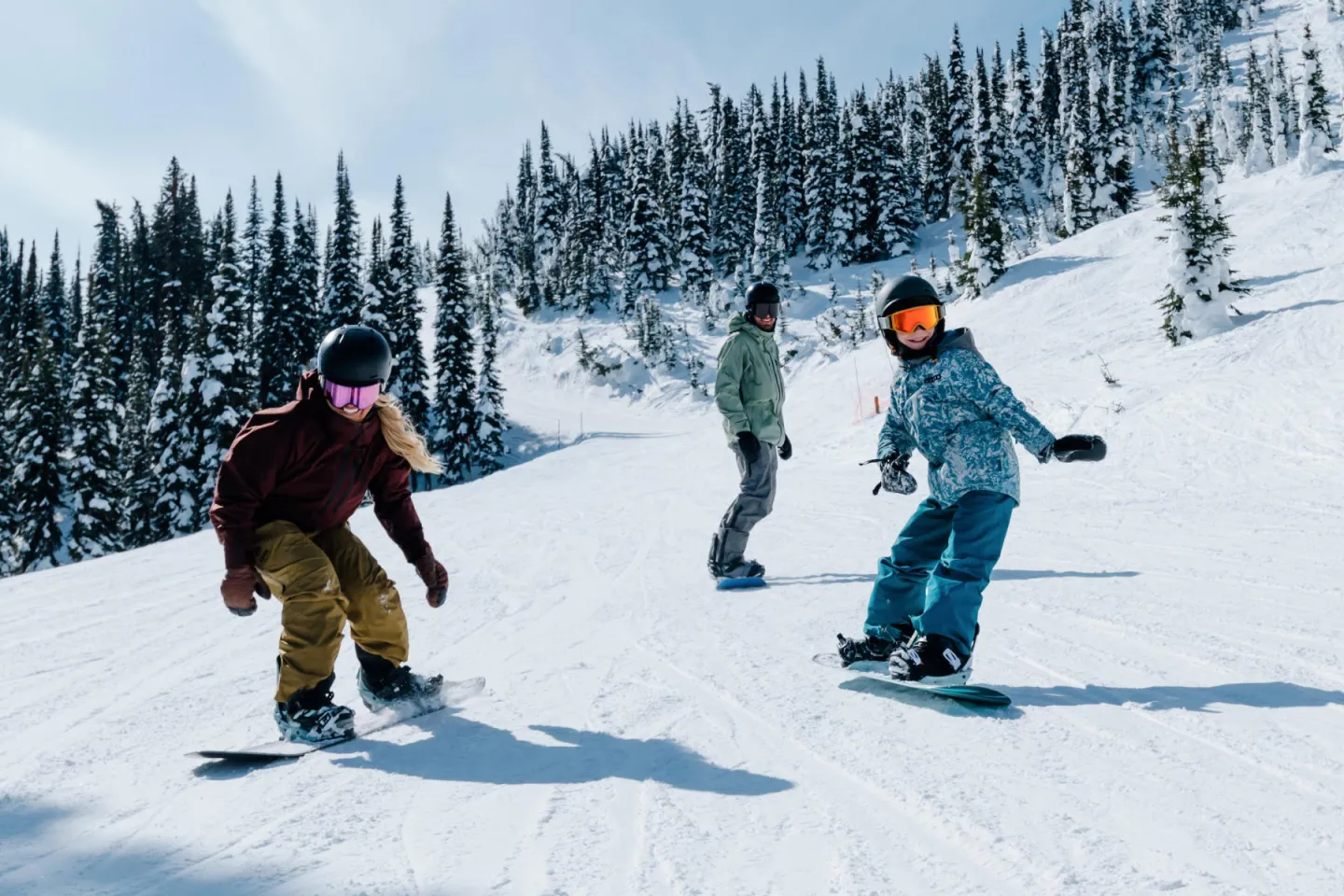 Three people snowboarding downhill on a sunny day, surrounded by snowy trees.