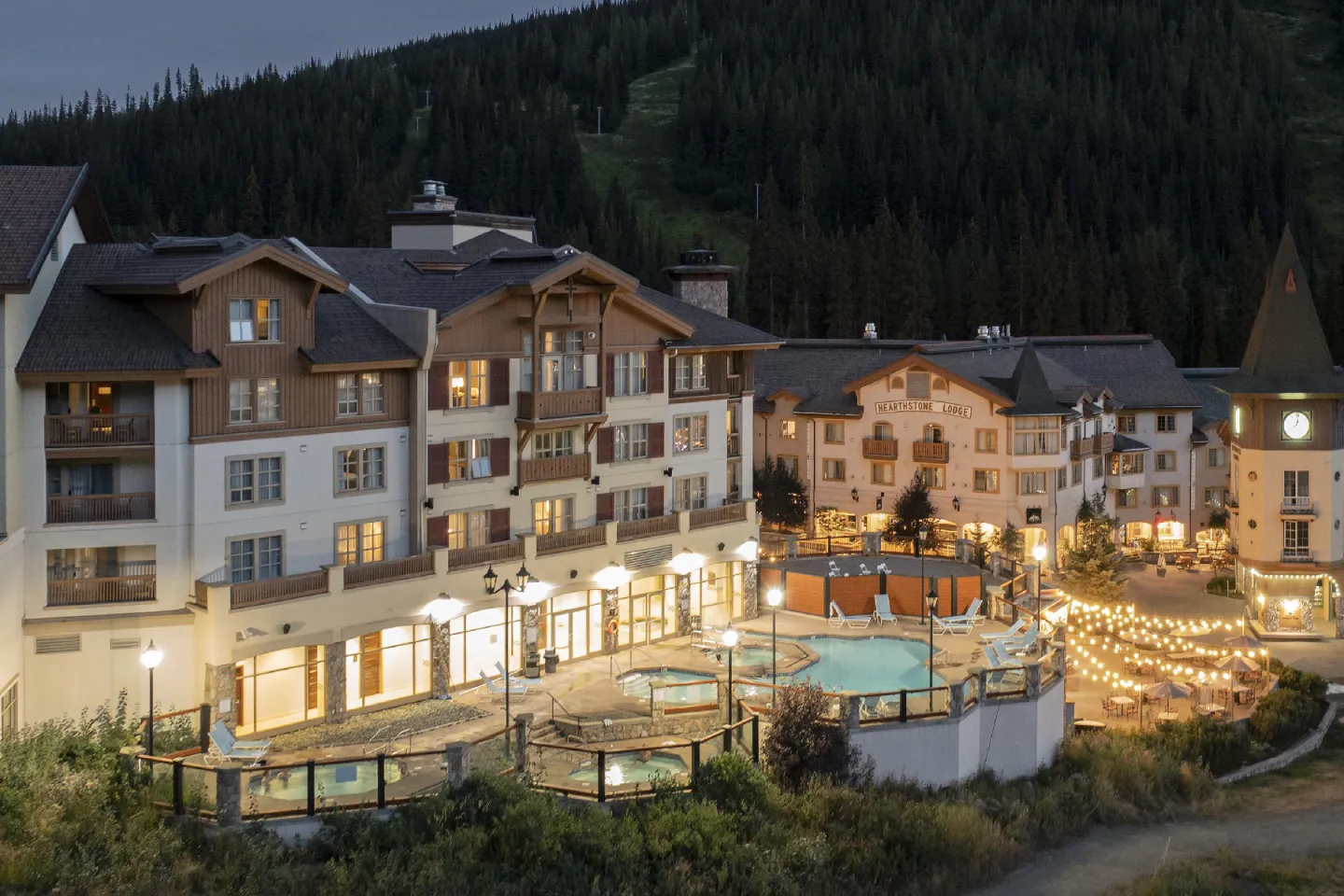 Ski resort at dusk with lit buildings, a pool, and surrounding mountains.