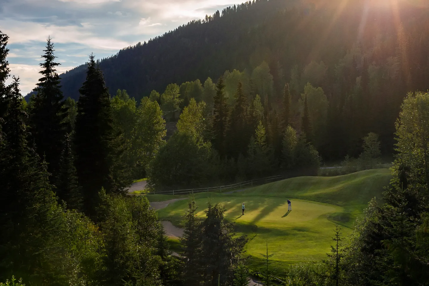 Golfers on a lush green course, surrounded by tall trees and mountains at sunset.
