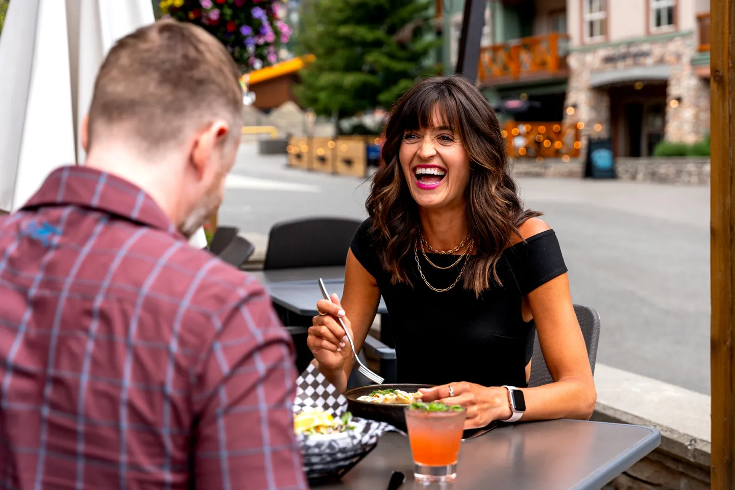 Woman laughing while dining outdoors with a man in a casual setting.