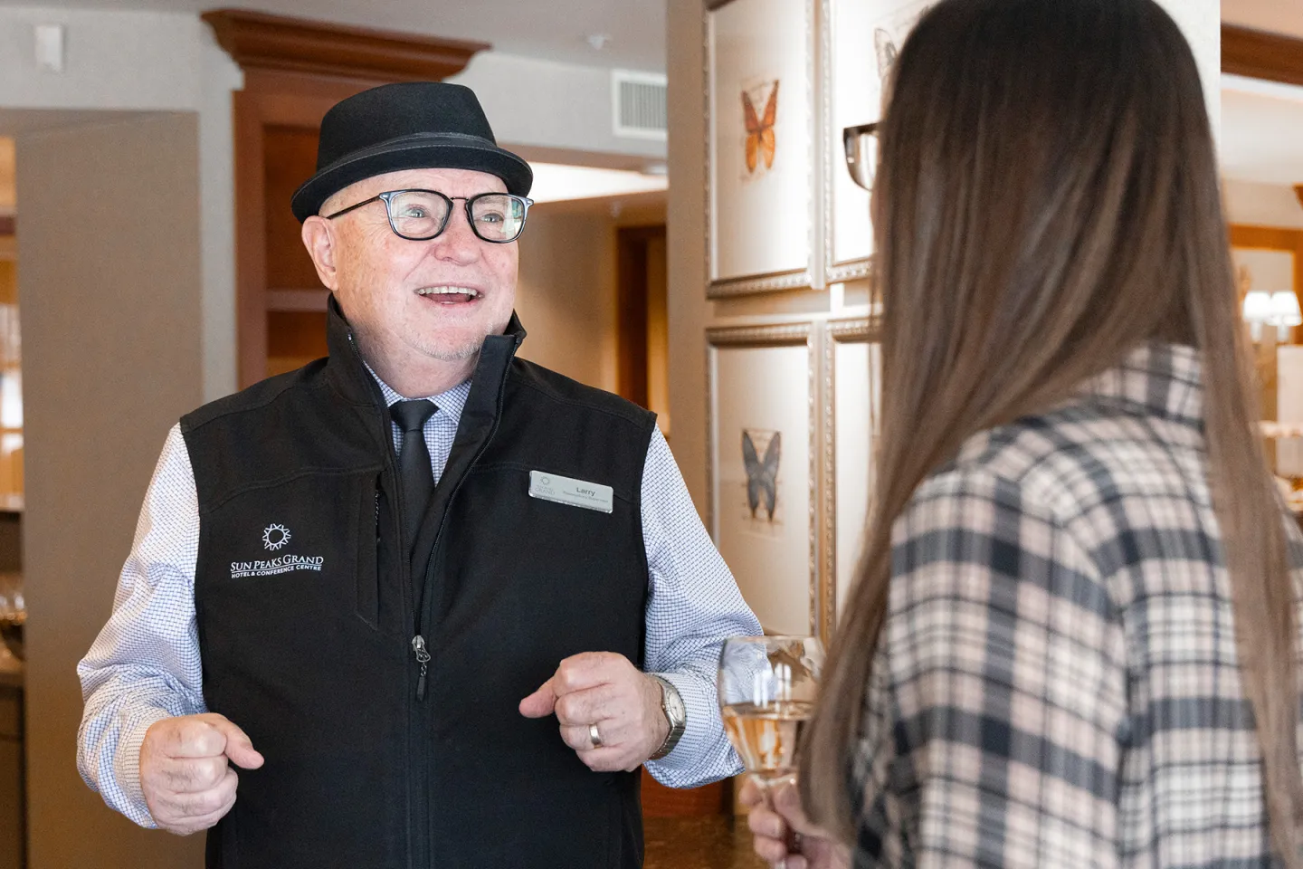 Man in black vest talks to woman in plaid jacket indoors.