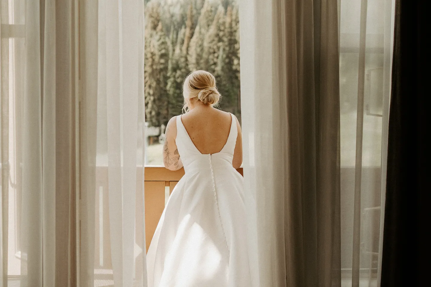 Woman in white dress standing on a balcony, viewed from behind, curtains framing her.