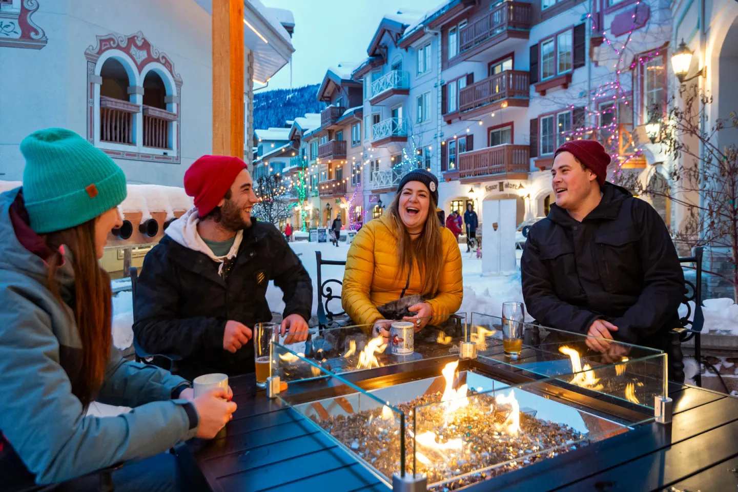 Friends laughing around an outdoor fire pit in a snowy village street.