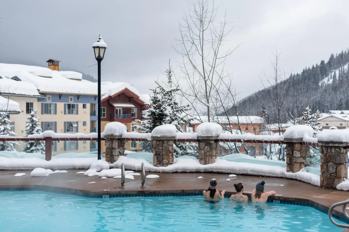 Outdoor pool with people swimming, surrounded by snowy buildings and trees.