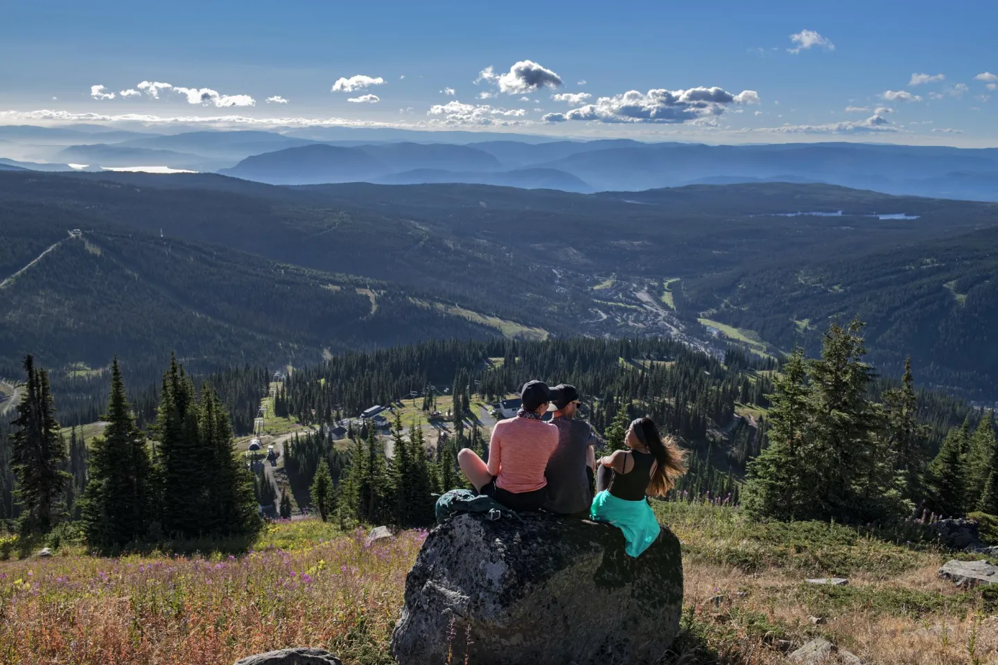 Three people sit on a rock overlooking a vast mountain landscape under a clear blue sky.