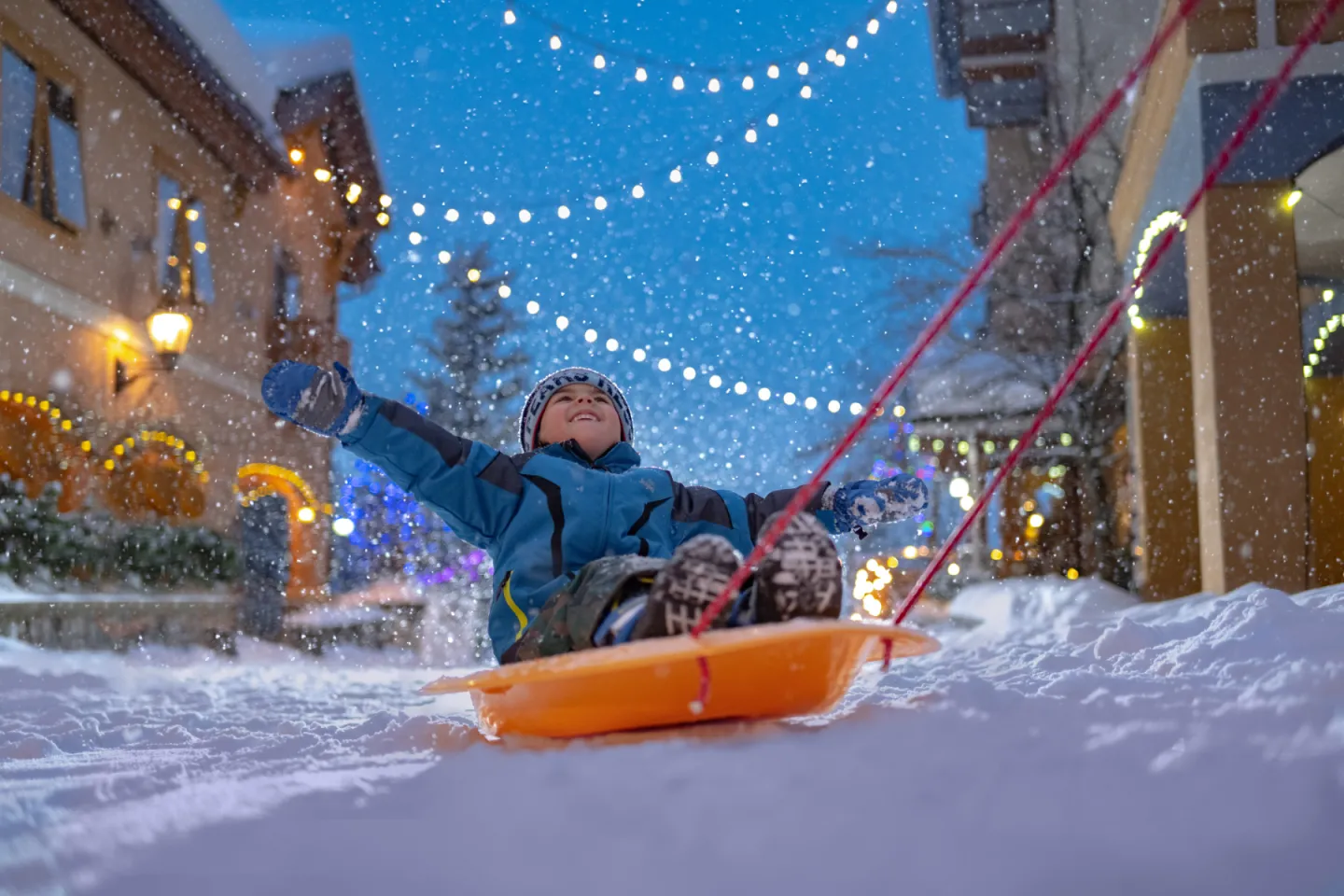 Child sledding joyfully in snowy town, under string lights.