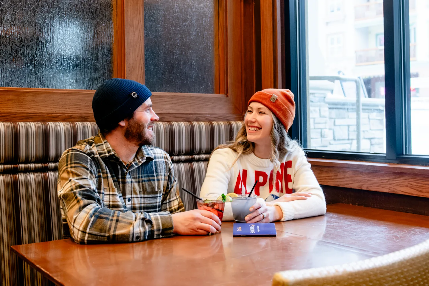 Man and woman in beanies smiling at each other in a cozy restaurant booth..