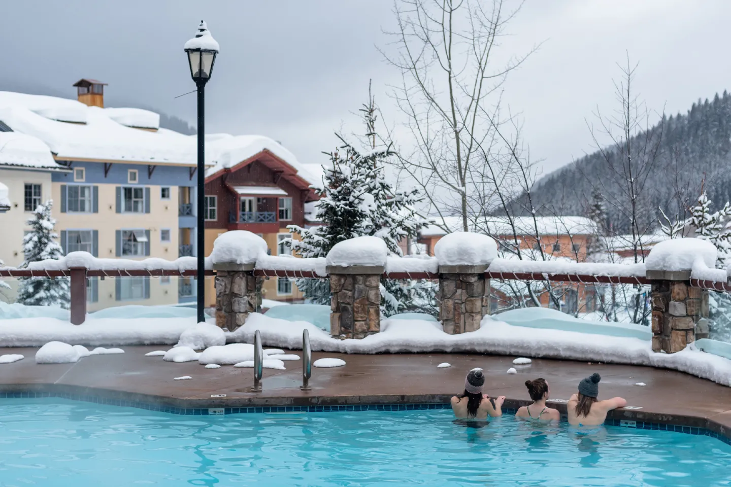 Three people in a snowy outdoor pool near chalet-style buildings.