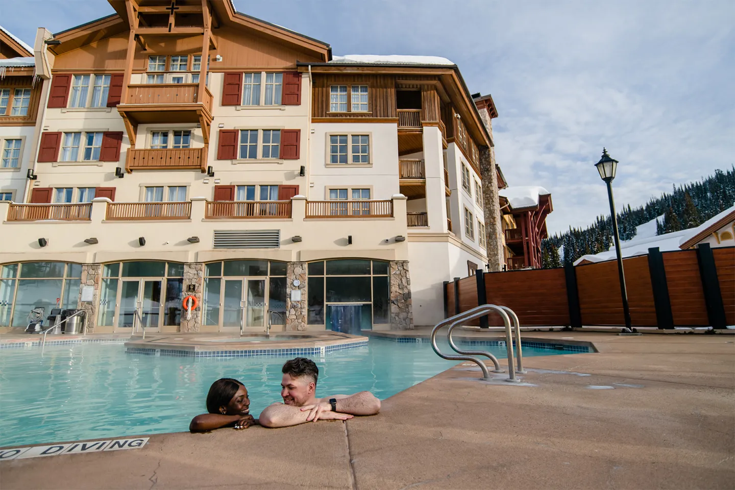 Couple relaxing in a pool near a large hotel, surrounded by snowy mountains.