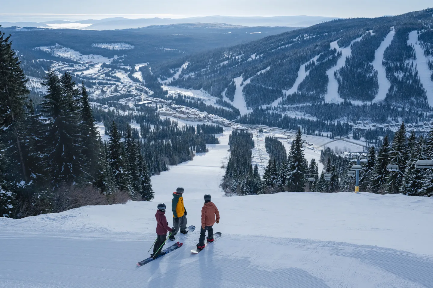 Three skiers on a snowy mountain slope with distant hills and pine trees.