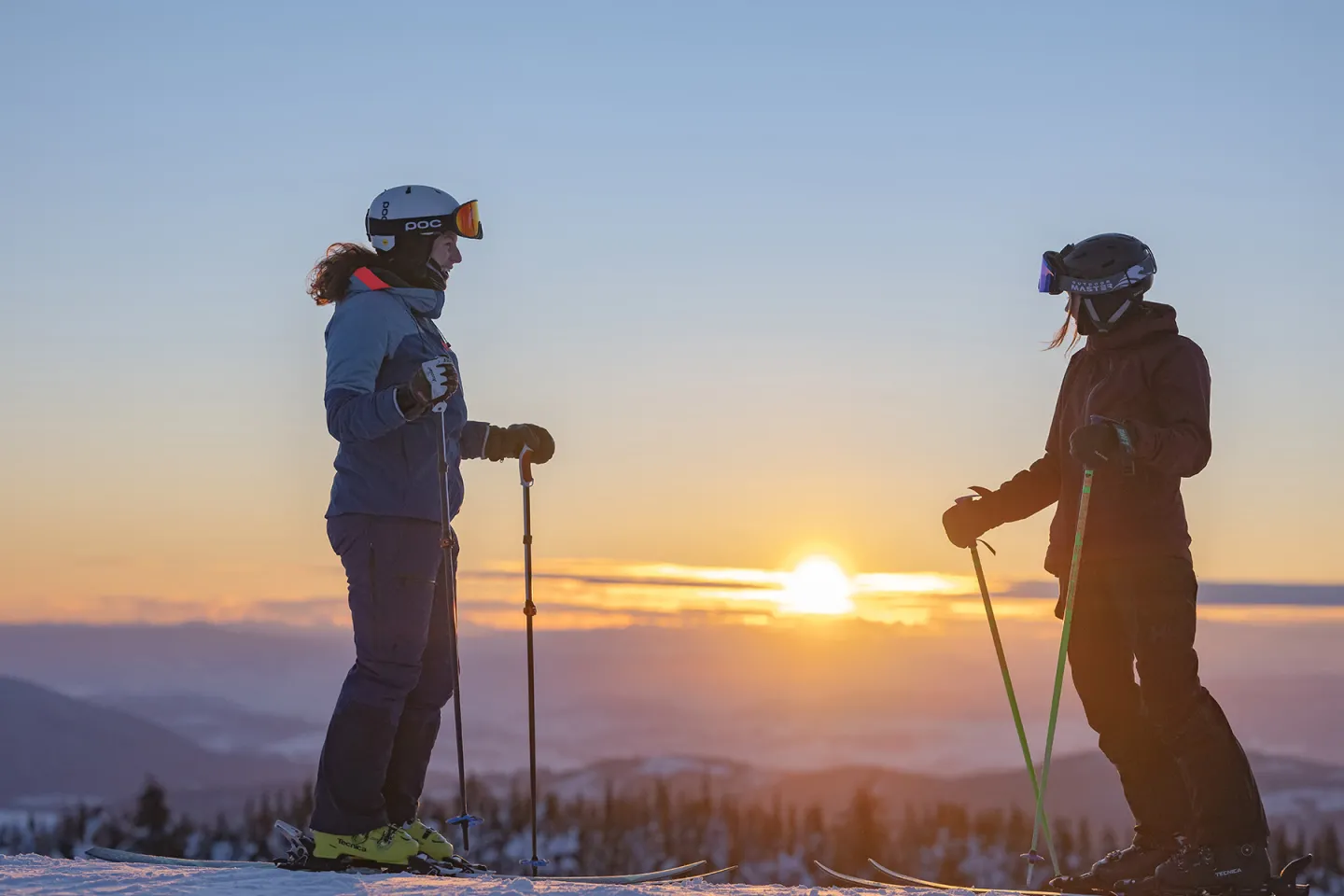 Two skiers stand facing each other at sunset on a snowy mountain.