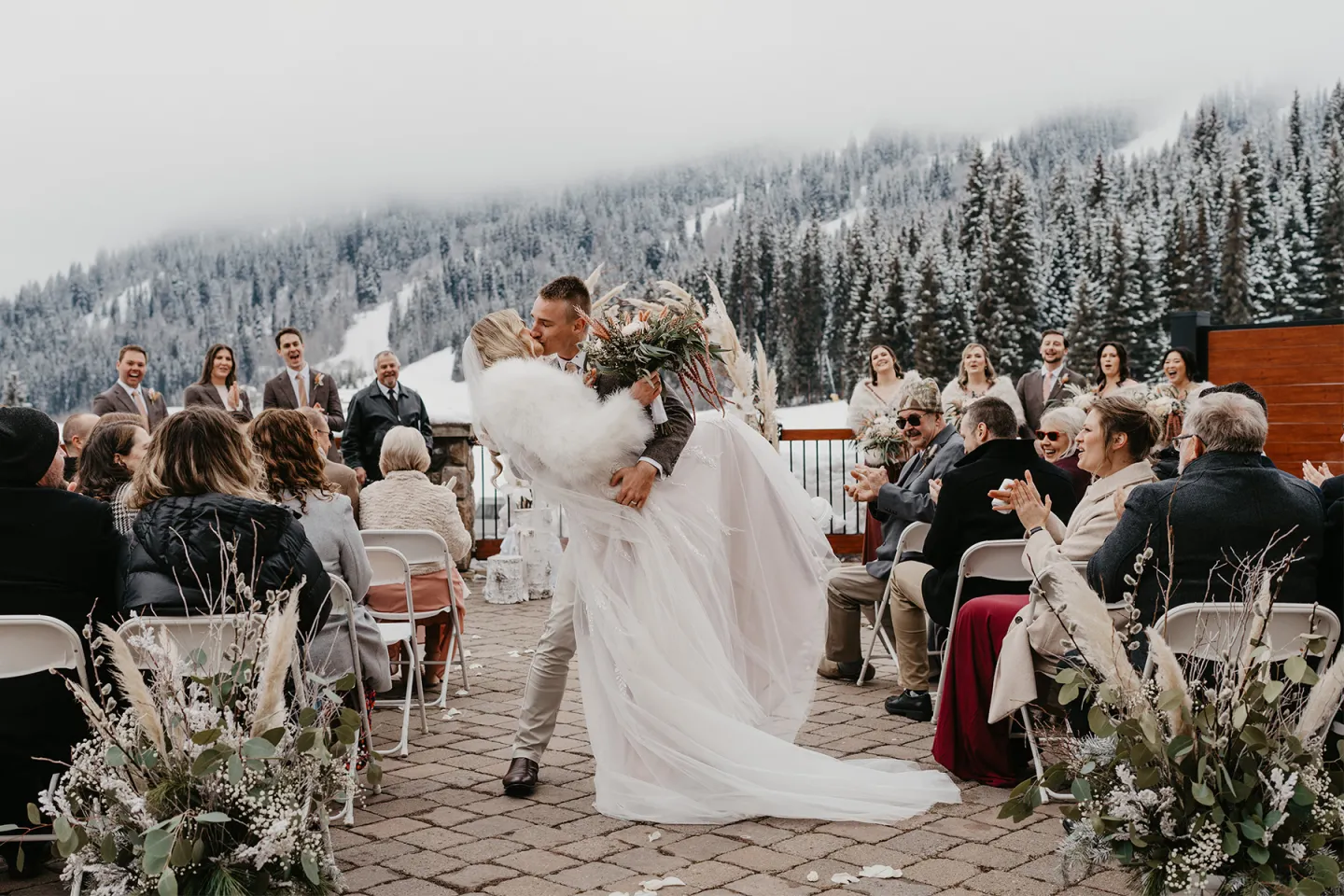 Groom kissing bride on snowy mountain patio with guests seated around.