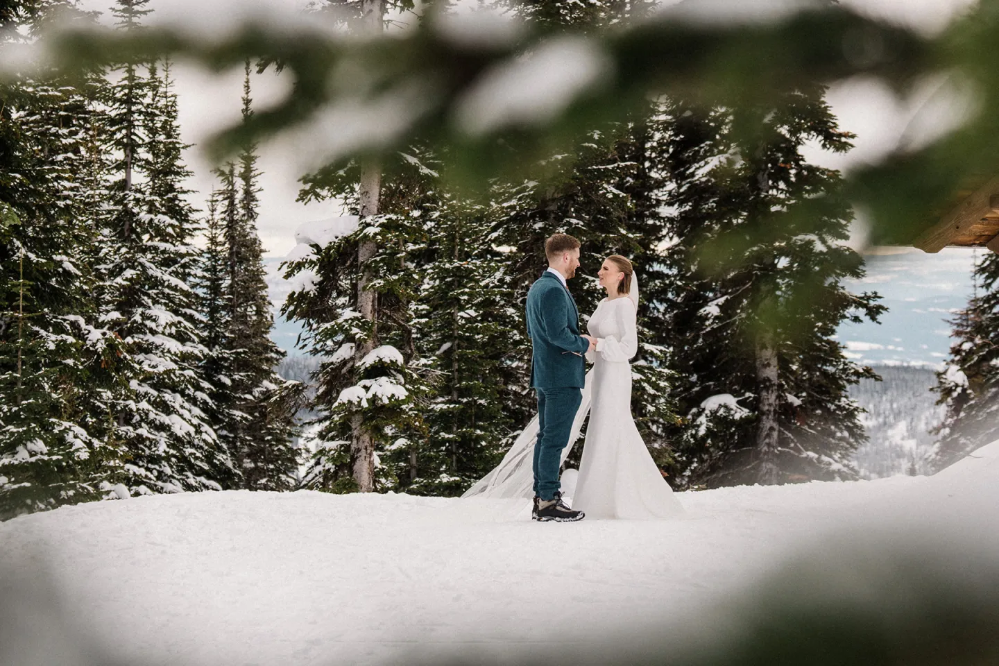 Couple in wedding attire stands in a snowy forest clearing.