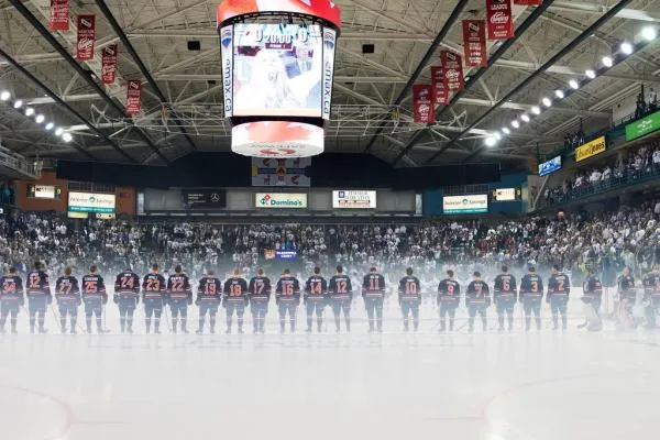 Hockey players lined up on an ice rink with a smoky backdrop and a large overhead screen.