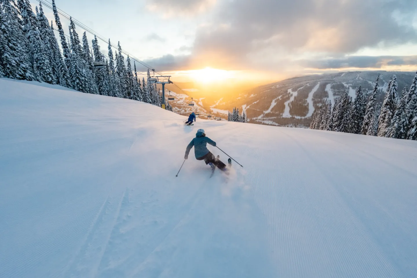 Skiers descending a snowy slope at sunrise with trees and mountains in the background.