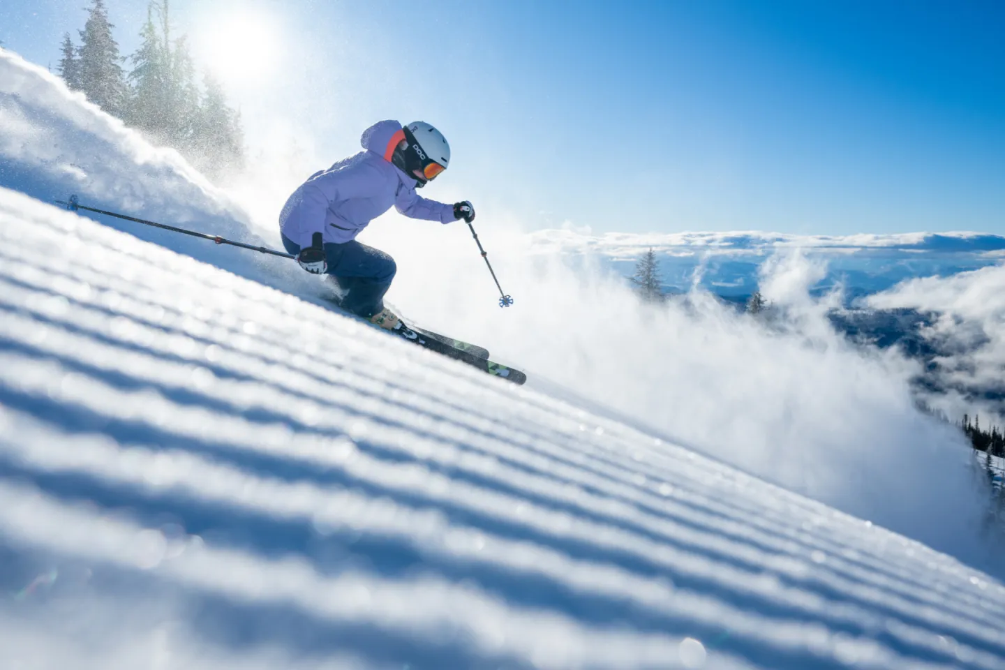 Skier in action on a sunny, snowy slope with trees and blue sky.
