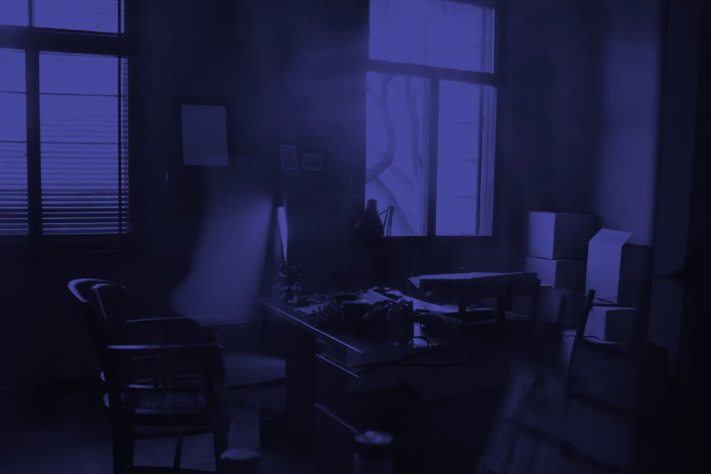 Dimly lit office with desk, chair, and stacked boxes, tinted in blue.