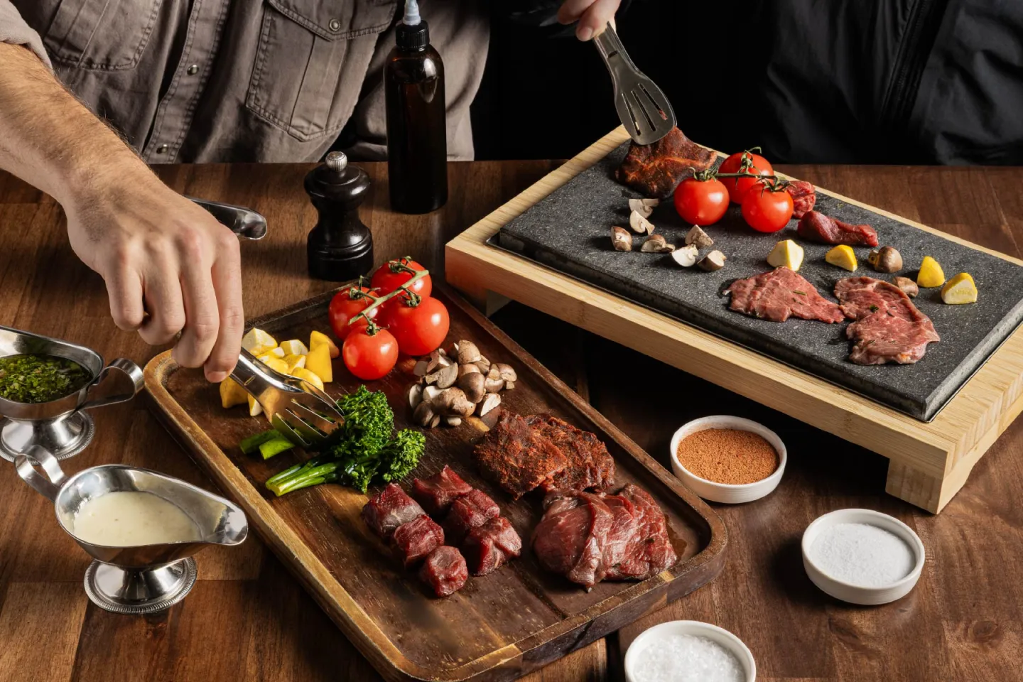 Steak on a hot stone with vegetables, sauces, and side dishes on a wooden table.