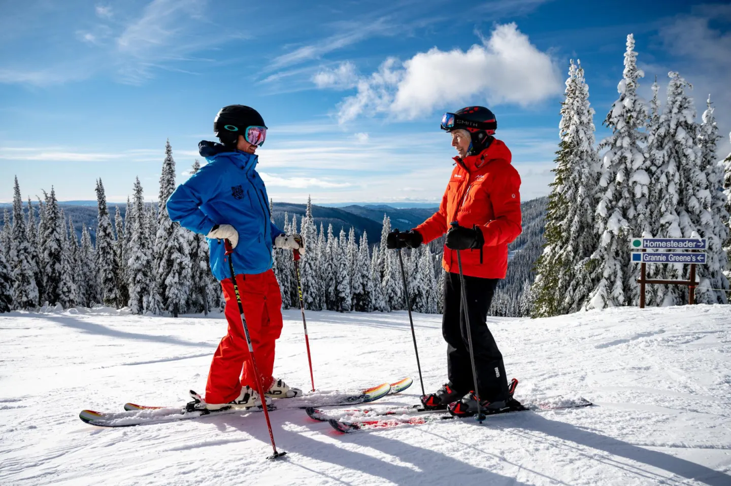 Nancy Greene and another skier in colorful gear talking on a snowy slope, surrounded by snow-covered trees.