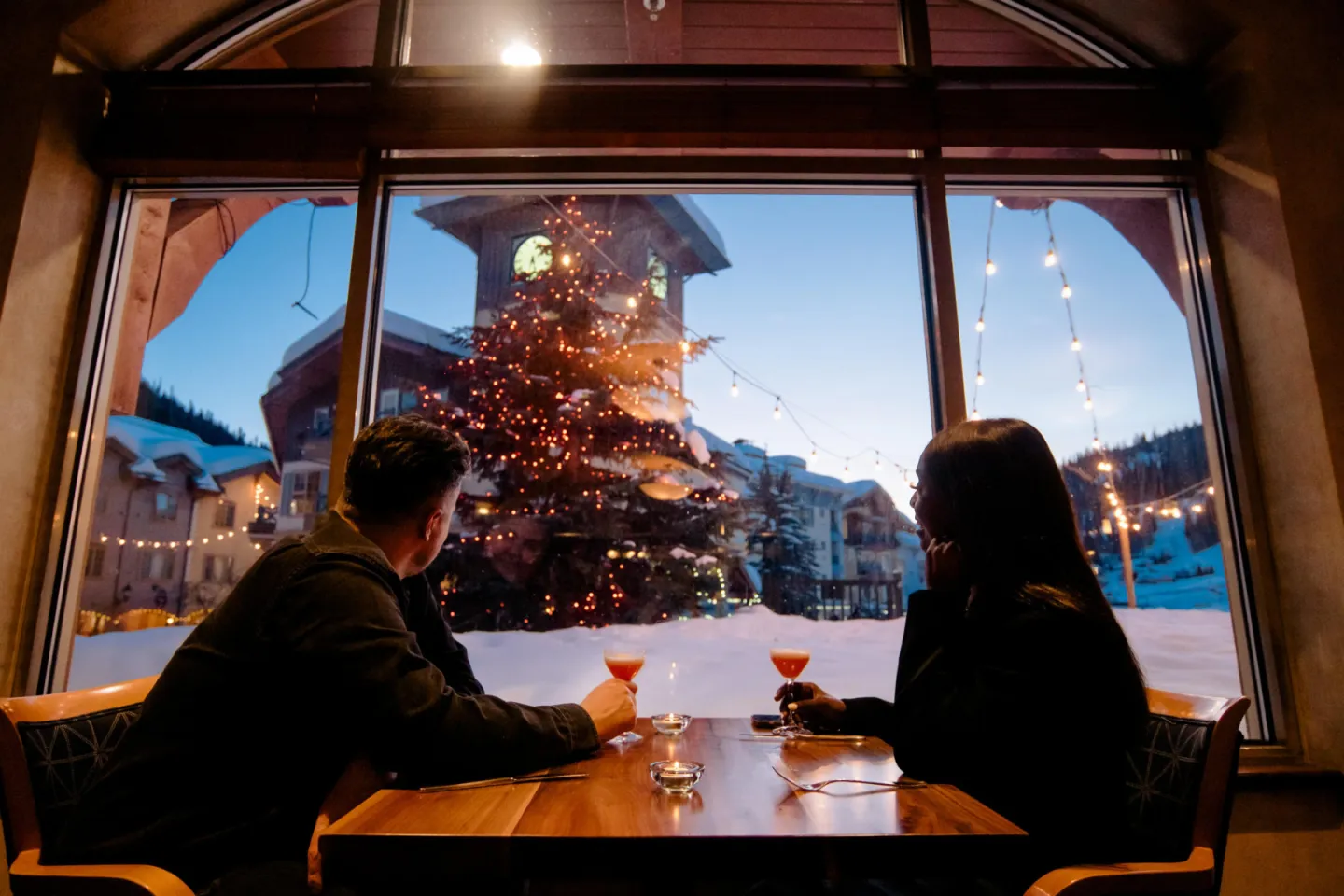 Couple enjoys drinks by a window with snowy village and Christmas tree.