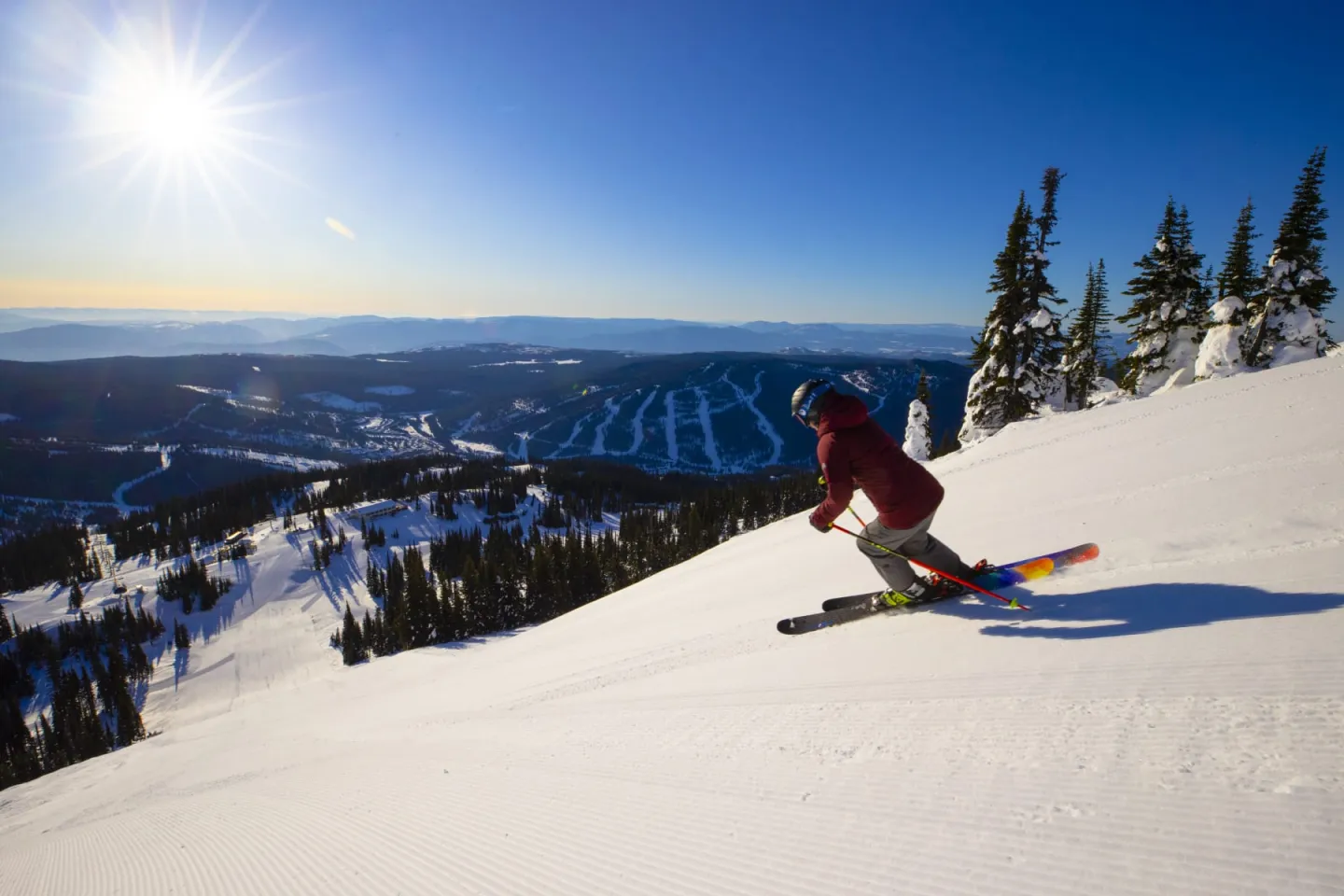 Skier descending a snowy slope under a bright sun, with a mountainous landscape in the background.