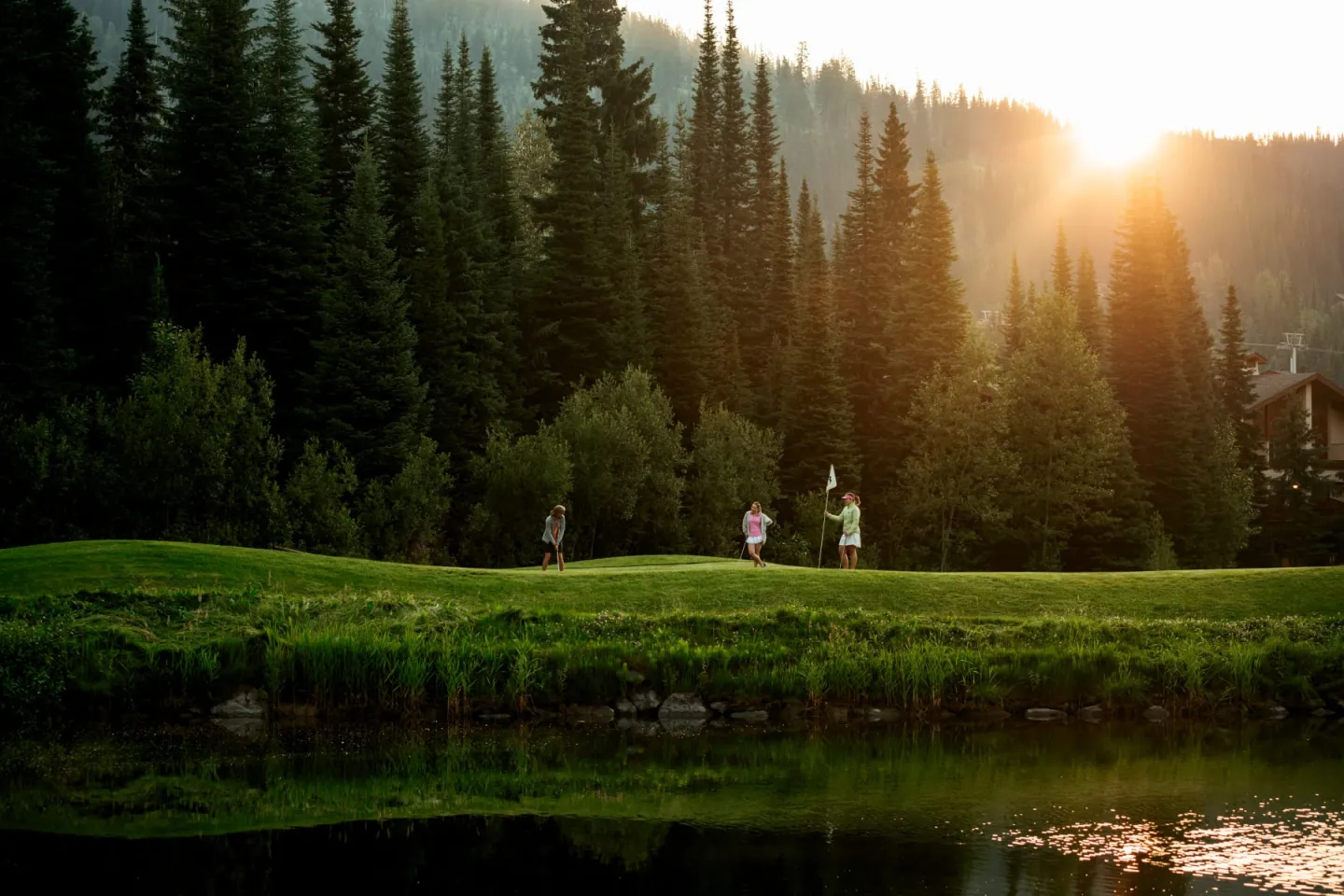 Sunset over a forest with people walking on a golf green by a pond.