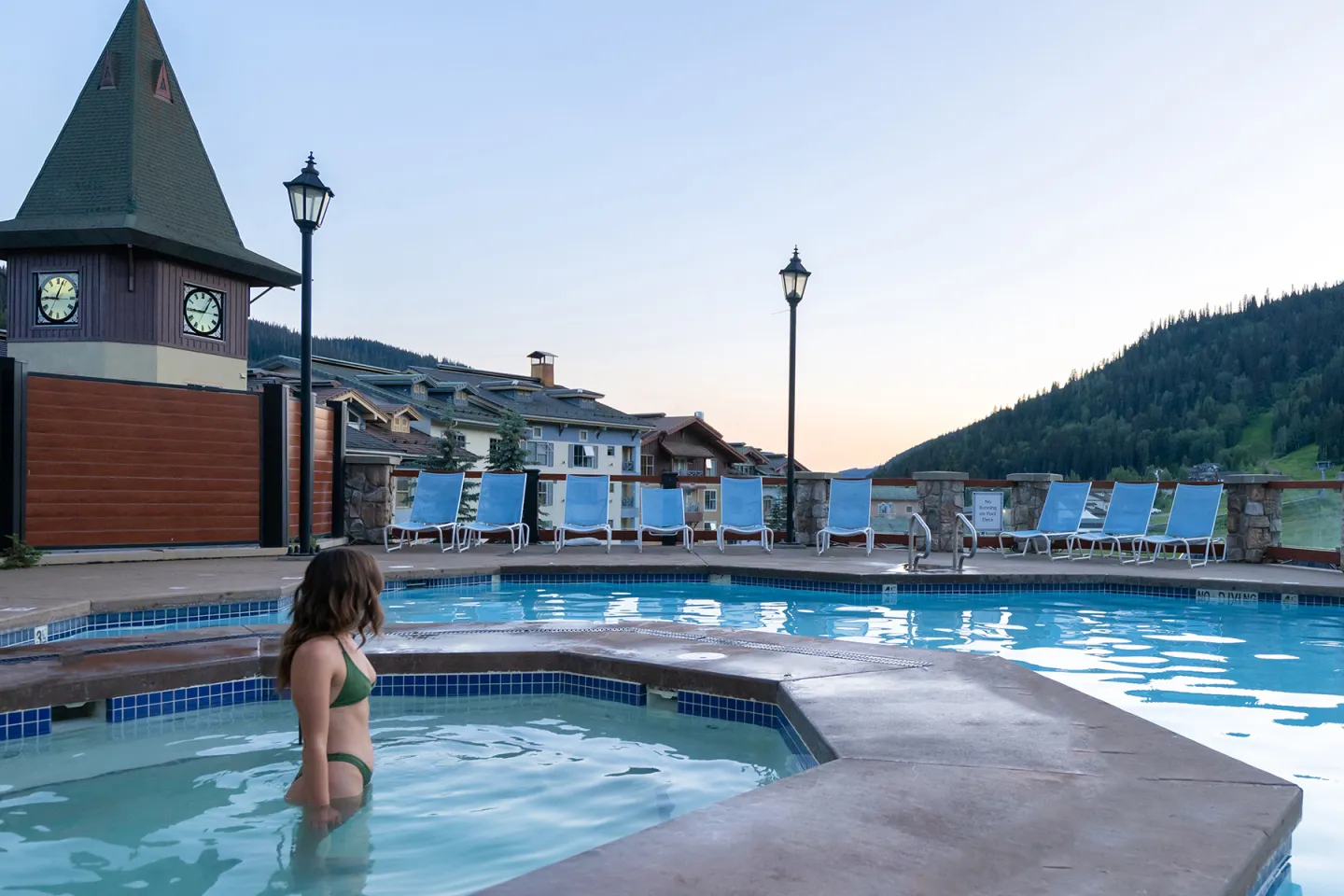 Woman in a hot tub by a pool at dusk, mountains in the background.