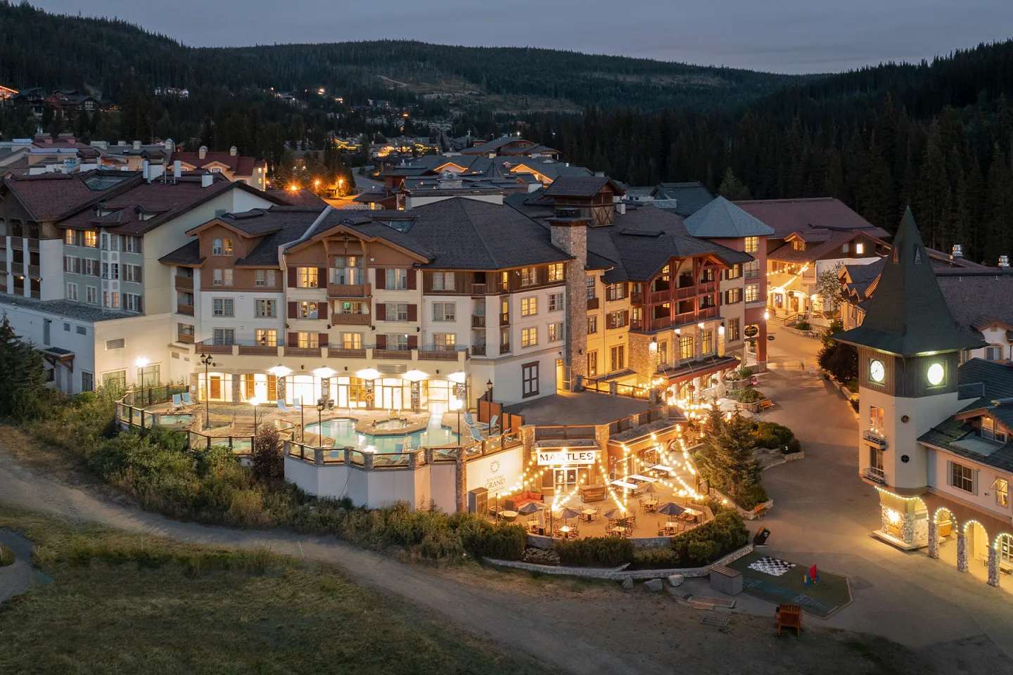 Mountain resort at dusk, warmly lit buildings, surrounded by forested hills.