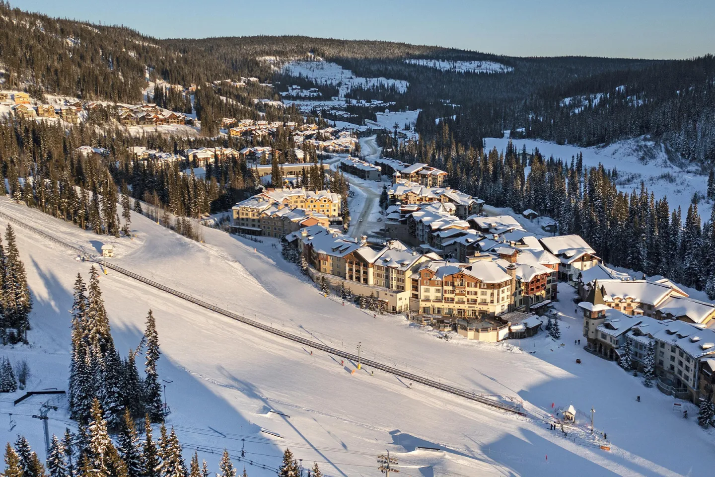 Snowy mountain village at sunrise, buildings nestled among pine trees.