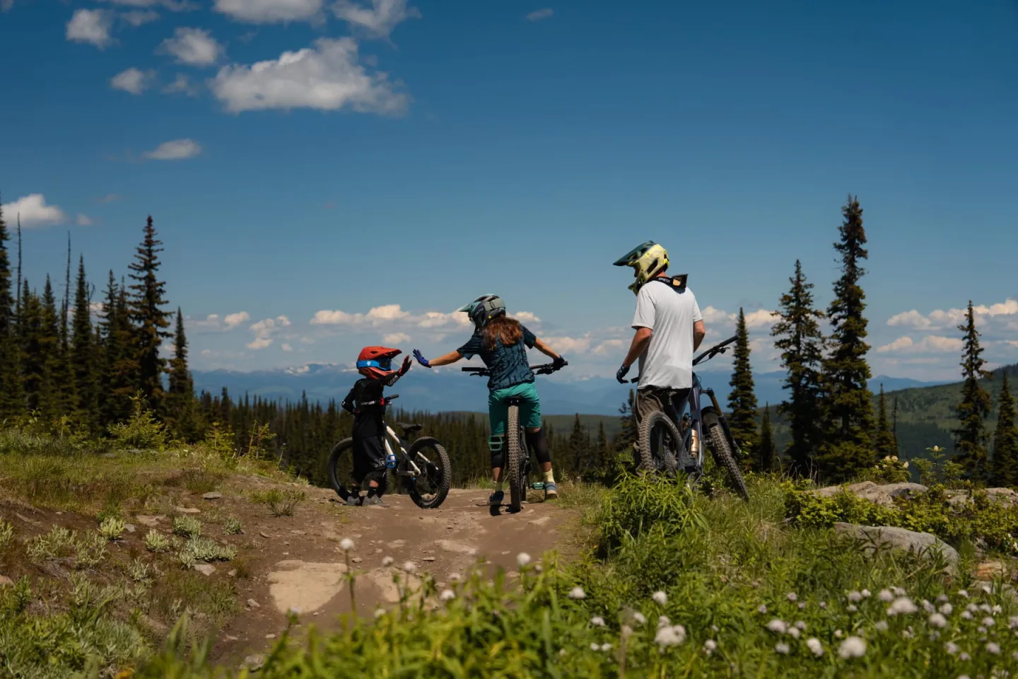 Bikers on a scenic trail surrounded by trees and mountains under a clear blue sky.