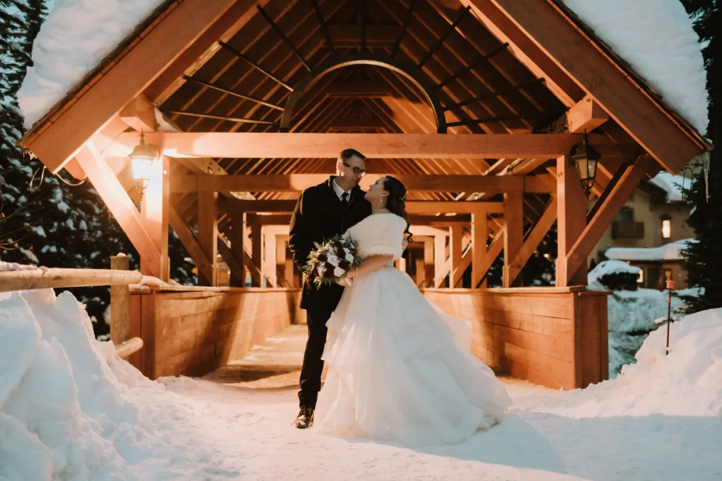 Bride and groom embrace under a snowy wooden archway at night.