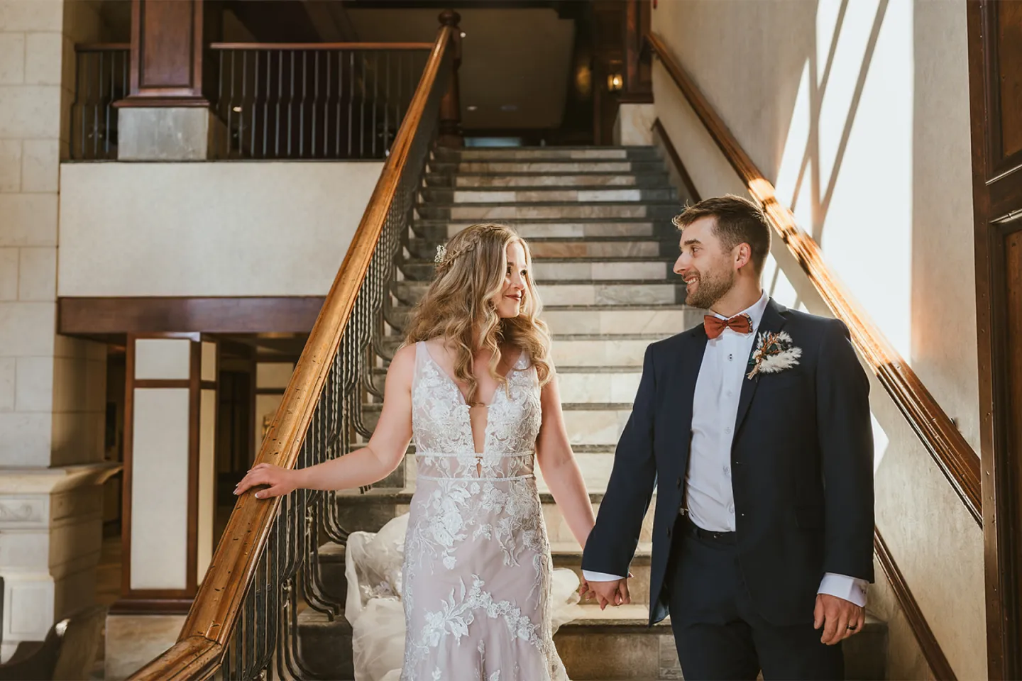 Bride and groom holding hands on a sunlit staircase, smiling at each other.