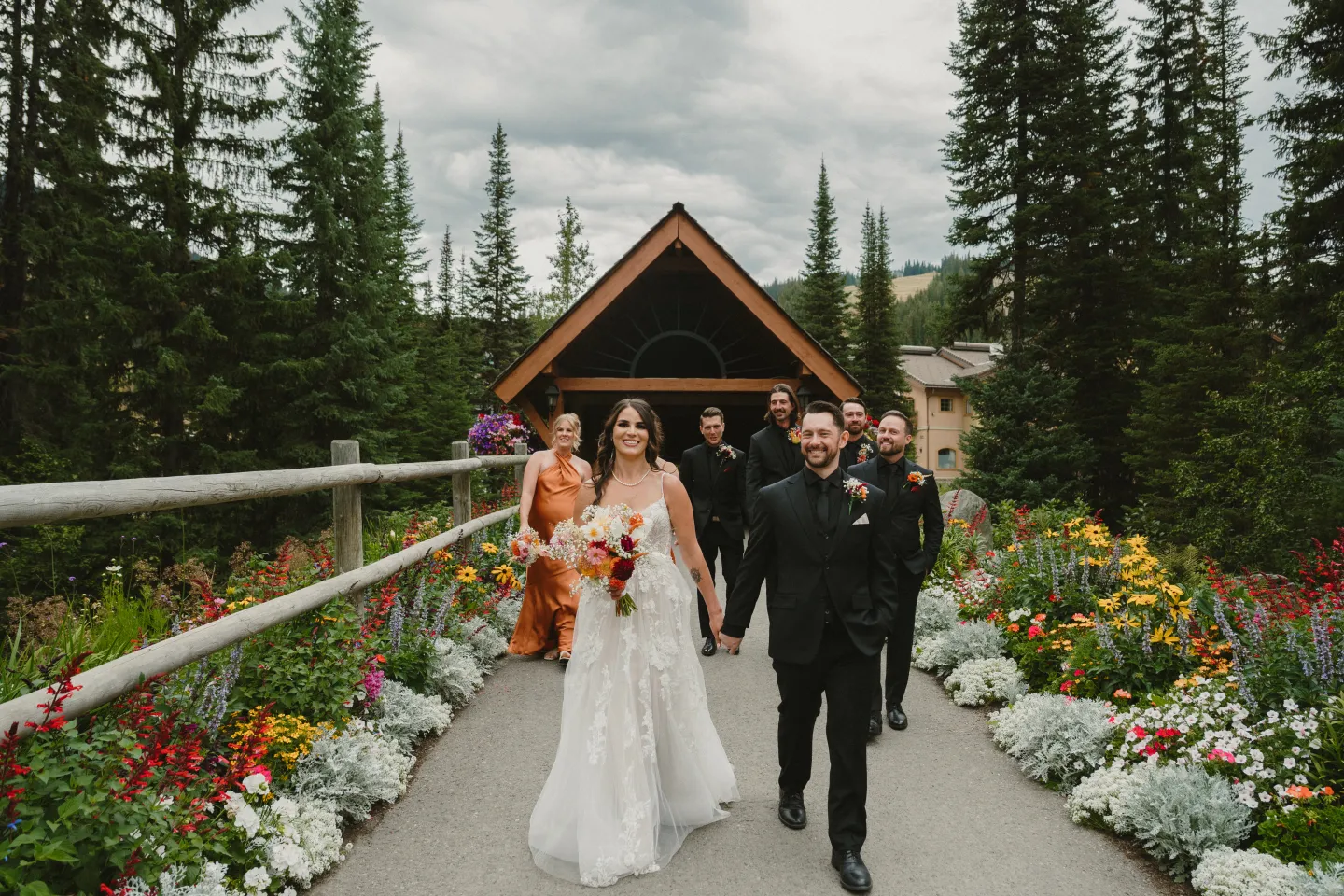 Bride and groom walk down flower-lined path, followed by wedding party in forest setting.
