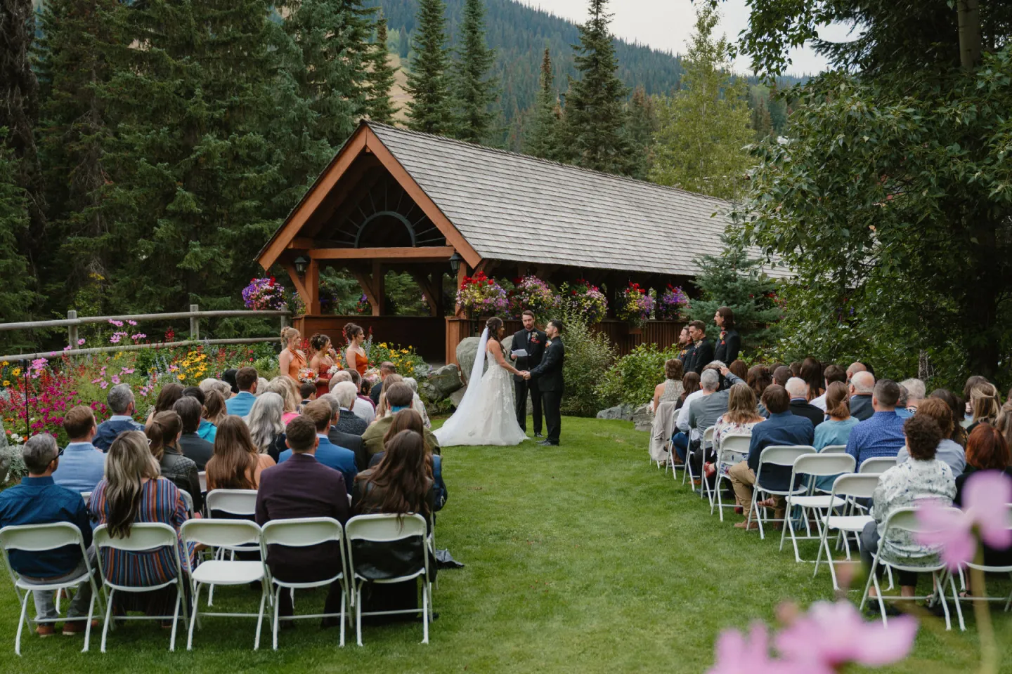 Wedding ceremony outdoors with guests seated, surrounded by trees and flowers.