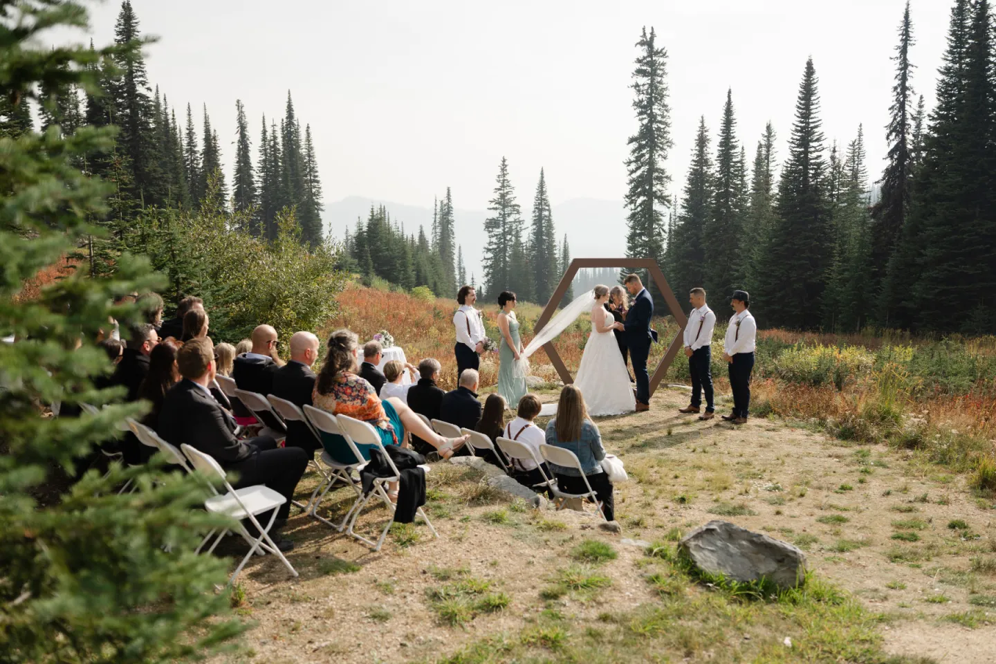 Outdoor wedding in forest with guests seated, bride and groom at wooden arch.