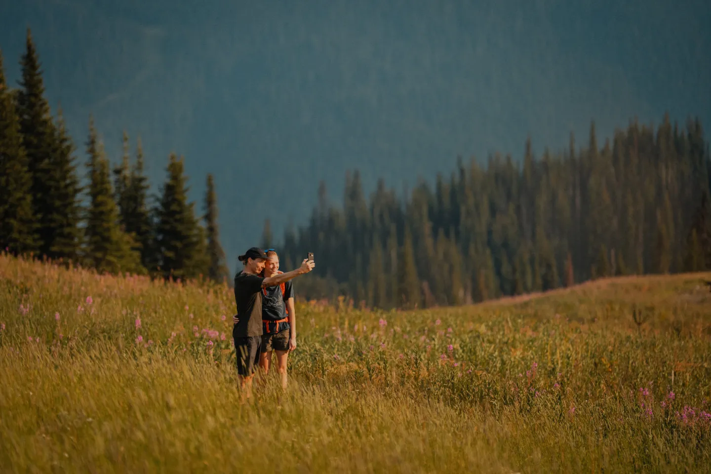 Two people taking a selfie in a wildflower meadow with a forested mountain backdrop.