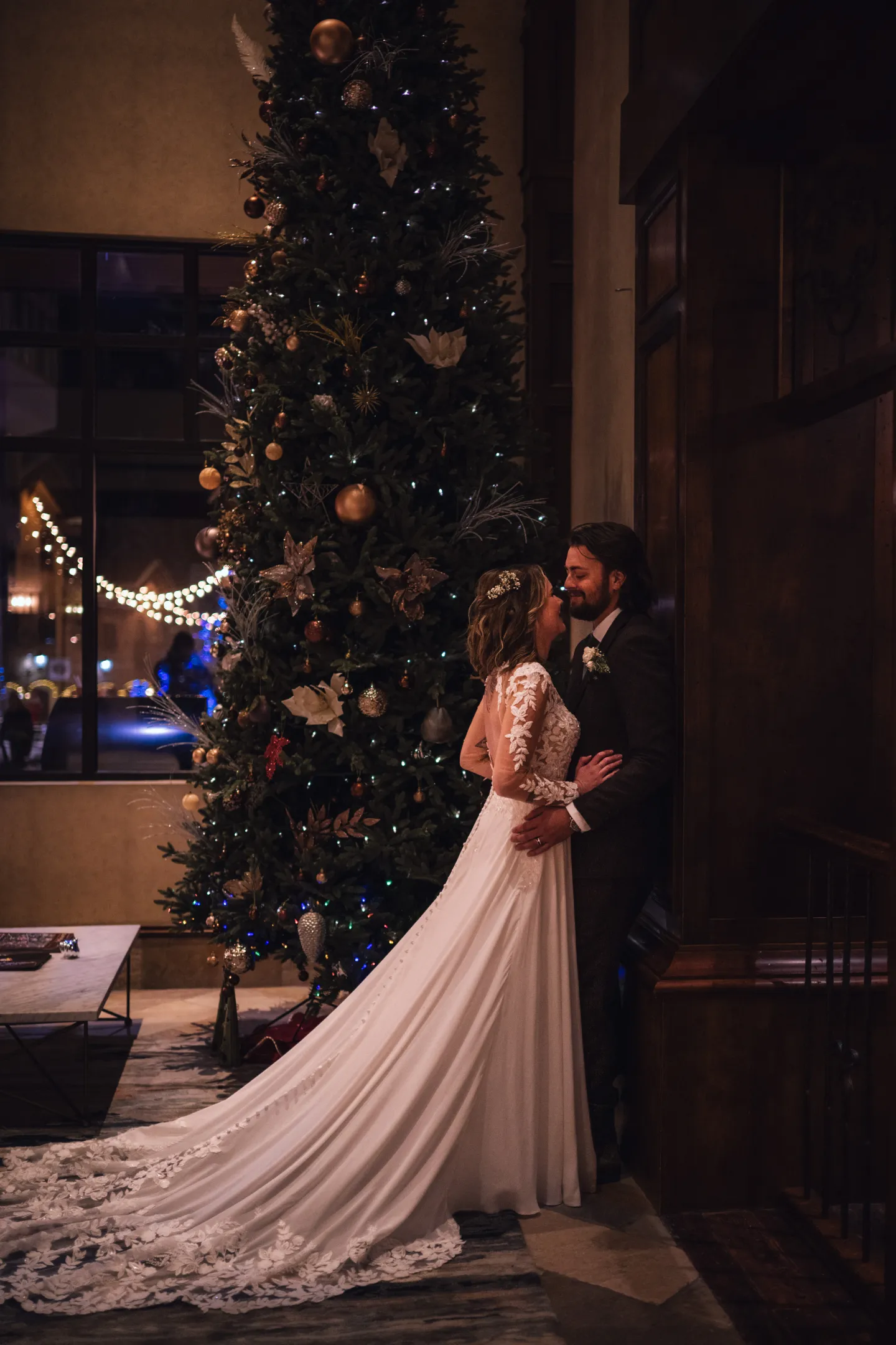 Bride and groom lean against a wall near a decorated Christmas tree.