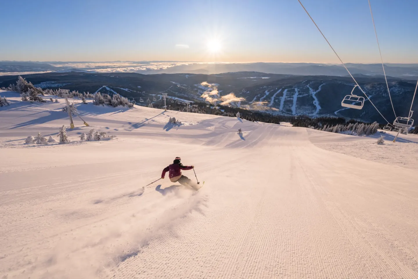 Skier descends snowy slope at sunrise with mountains in distance.