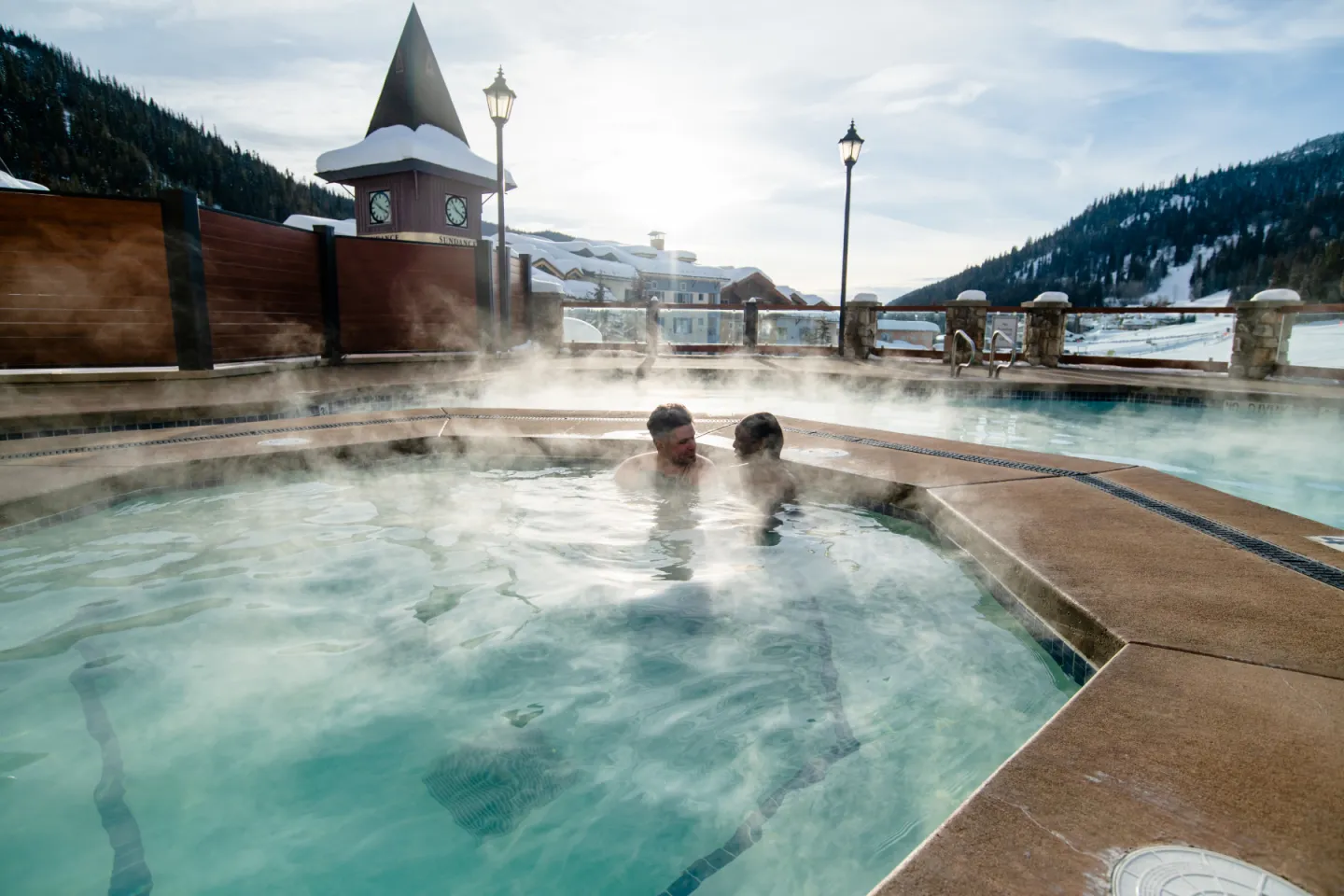 Two people soaking in a steaming outdoor pool beside snowy mountains.