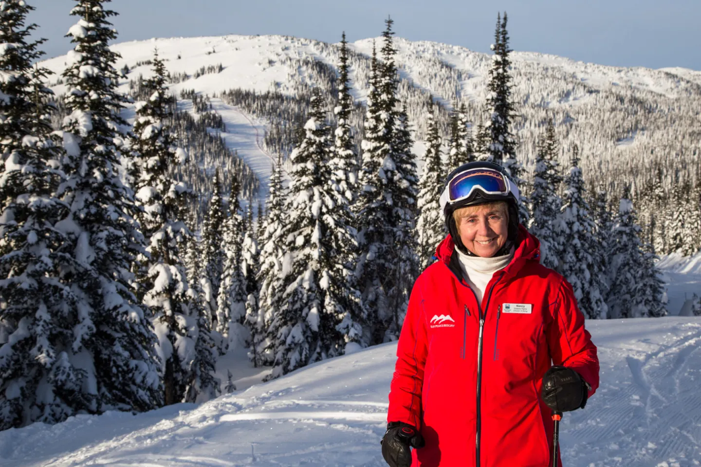 Smiling skier (Nancy Greene) in a red jacket stands on a snowy mountain trail.