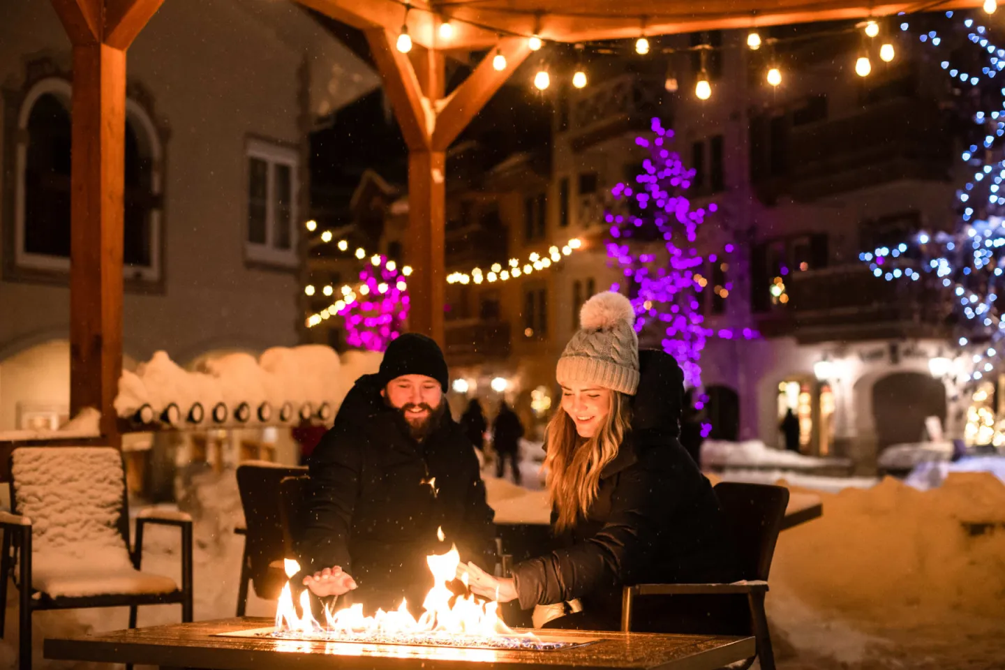 Two people gathered around a fire pit at a snowy, holiday-lit outdoor patio.